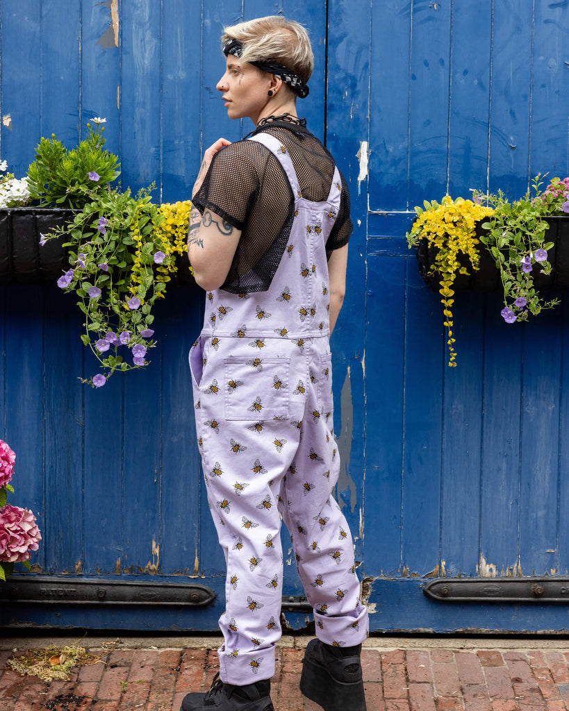 Jamie is wearing the Lavender bees stretch twill dungarees worn with a black fishnet top and platform boots, hem is rolled up. Model is facing away from the camera with hands in pockets and standing in front of a garage with cobbled floors and flowers.