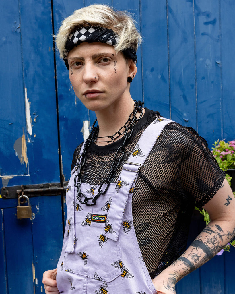 Jamie is wearing the Lavender bees stretch twill dungarees worn with a black fishnet top and platform boots, hem is rolled up. Model is standing with hands in pockets, facing the camera and standing in front of a garage with cobbled floors and flowers.