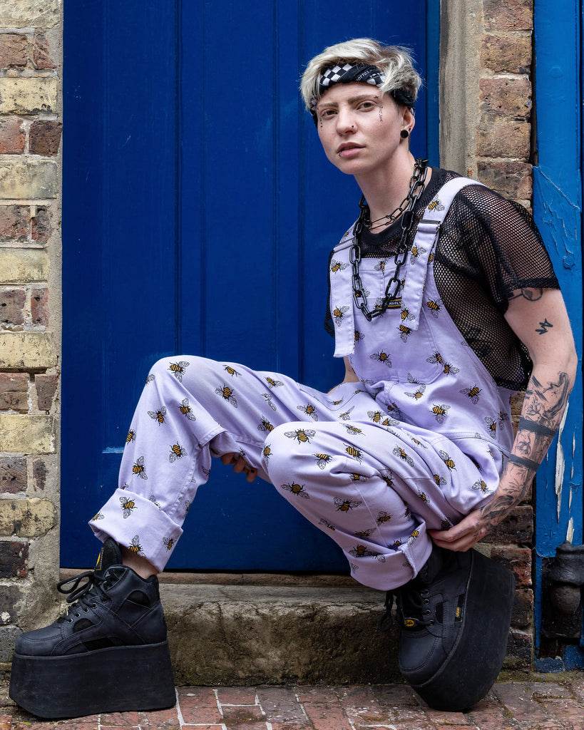 Jamie is wearing the Lavender bees stretch twill dungarees worn with a black fishnet top and platform boots, hem is rolled up. Model is crouched down, facing the camera and standing in front of a garage with cobbled floors 