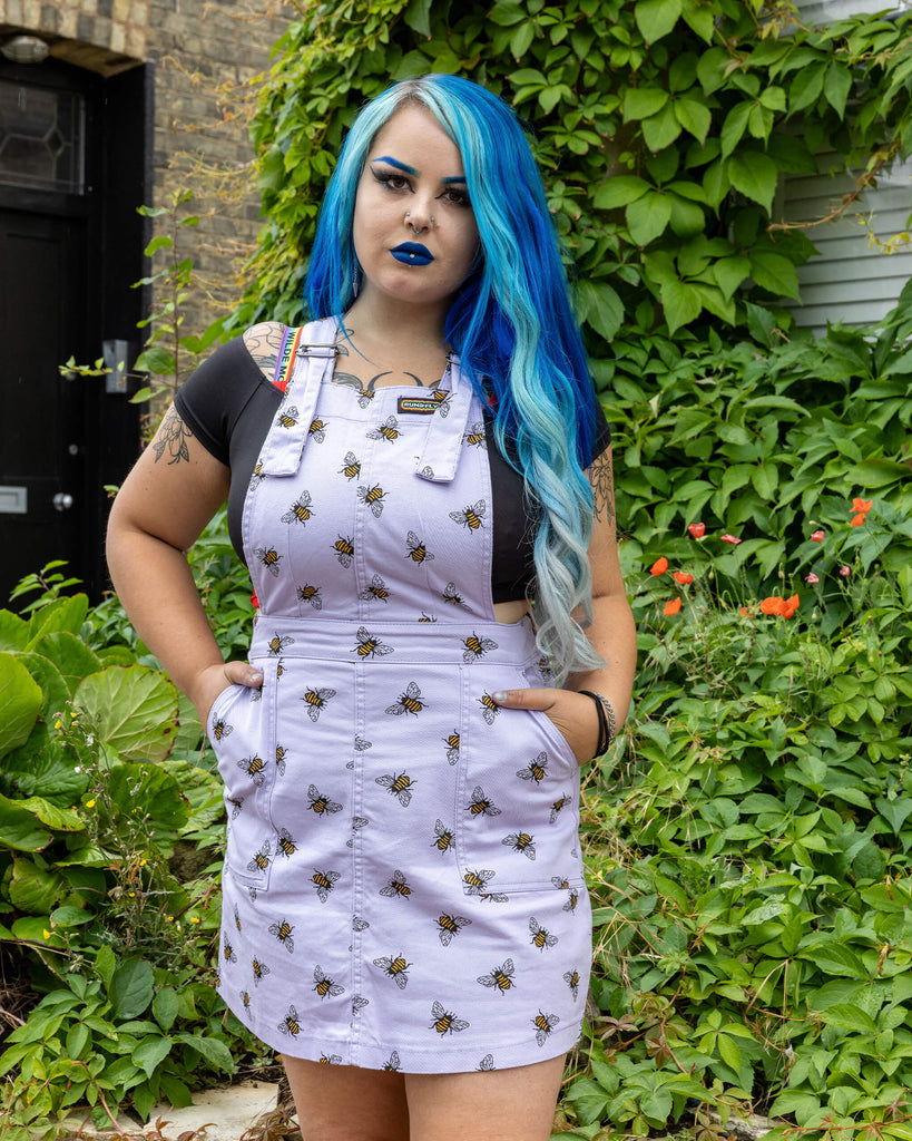Georgina is wearing the Lavender bees stretch twill pinafore dress worn with a black short sleeved top and pink jelly shoes, hem is rolled up. Model is standing with hands in pockets, facing the camera and standing in front of the garden of a brick house with cobbled floors and flowers.
