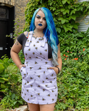 Georgina is wearing the Lavender bees stretch twill pinafore dress worn with a black short sleeved top and pink jelly shoes, hem is rolled up. Model is standing with hands in pockets, facing the camera and standing in front of the garden of a brick house with cobbled floors and flowers.