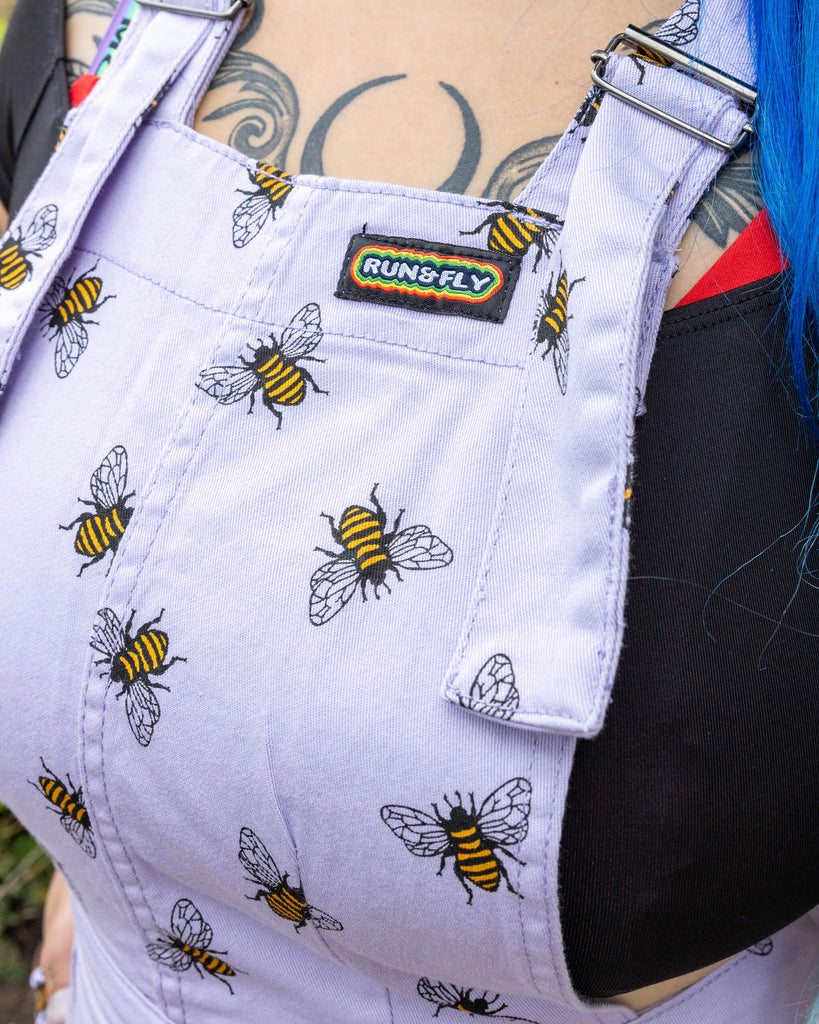 Closeup Georgina is wearing the Lavender bees stretch twill pinafore dress worn with a black short sleeved top and pink jelly shoes, hem is rolled up. Model is standing with hands in pockets, facing the camera and standing in front of the garden of a brick house with cobbled floors and flowers.