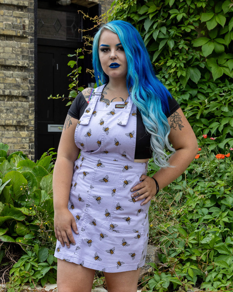 Georgina is wearing the Lavender bees stretch twill pinafore dress worn with a black short sleeved top. Model is standing with hands in pockets, facing the camera and standing in front of the garden of a brick house with cobbled floors and flowers.