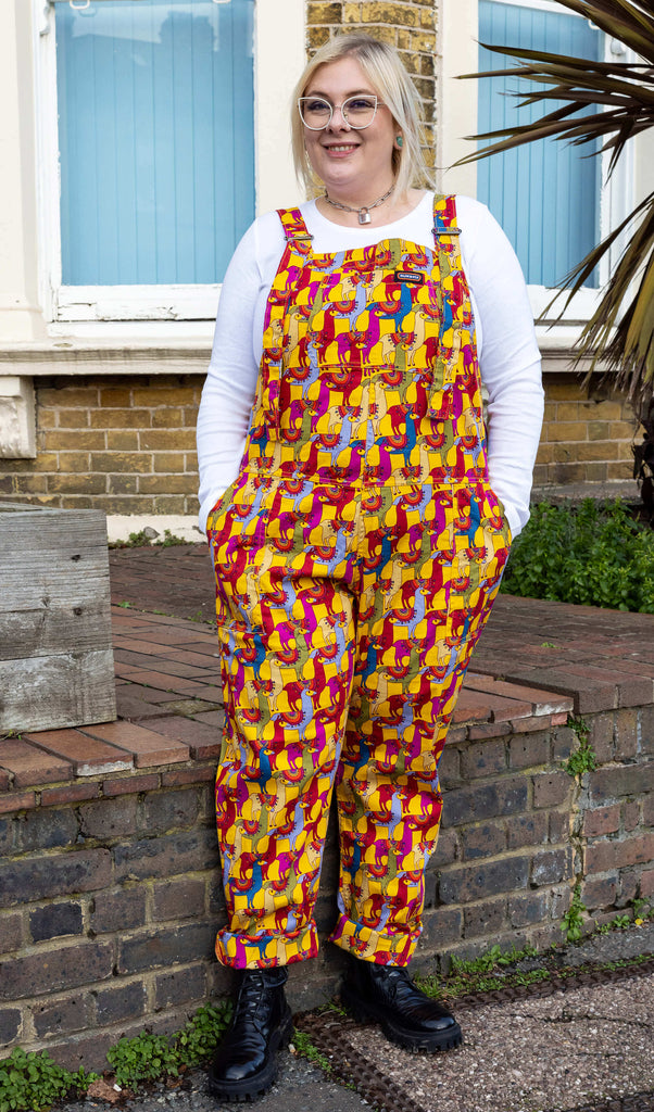 blonde haired female model outdoors wearing llama dungarees and a white long sleeved top with black boots smiling at camera