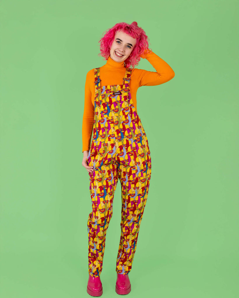 A model with vibrant pink curly hair is wearing bright yellow dungarees with a colourful llama print from Run & Fly. The dungarees are paired with a bold orange turtleneck and pink platform boots. The playful, eye-catching design features llamas in shades of red, blue, and purple. Run & Fly is a small, ethical, slow fashion company.