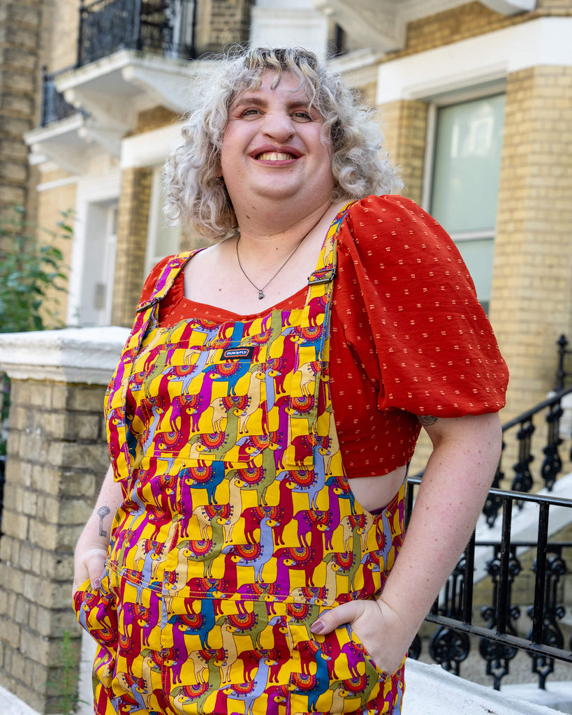 A close-up of a smiling model with curly blonde hair, wearing yellow llama print dungarees by Run & Fly. The dungarees feature a vibrant design with red, blue, and purple llamas. Paired with a red puff-sleeve top, the look is fun, colourful, and casual. Run & Fly is a small, ethical, slow fashion company.