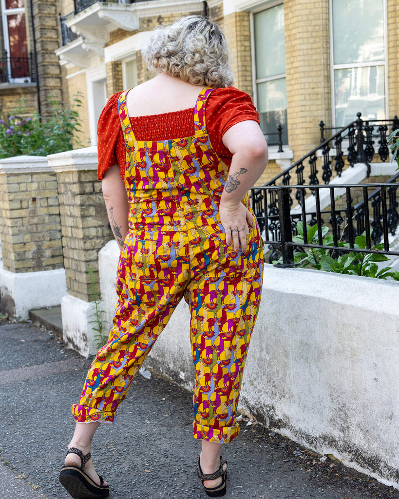 Back view of a model with curly blonde hair wearing yellow llama print dungarees from Run & Fly. The dungarees feature colourful llamas in red, purple, and blue. The model pairs them with a red puff-sleeve top and sandals, showcasing a casual and playful style. Run & Fly is a small, ethical, slow fashion company.