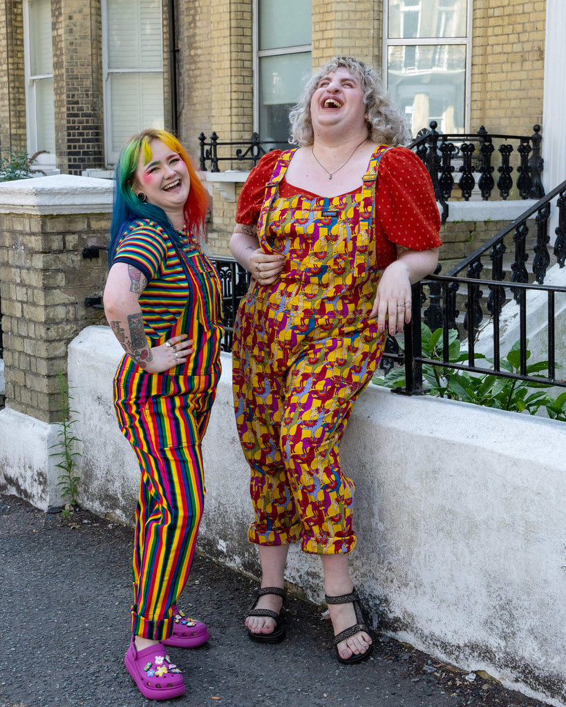 Two models are laughing and posing together outdoors. One wears rainbow-striped dungarees with pink Crocs, and the other wears yellow dungarees with a llama print by Run & Fly, paired with a red puff-sleeve top and sandals. The vibrant outfits create a joyful, playful vibe. Run & Fly is a small, ethical, slow fashion company.