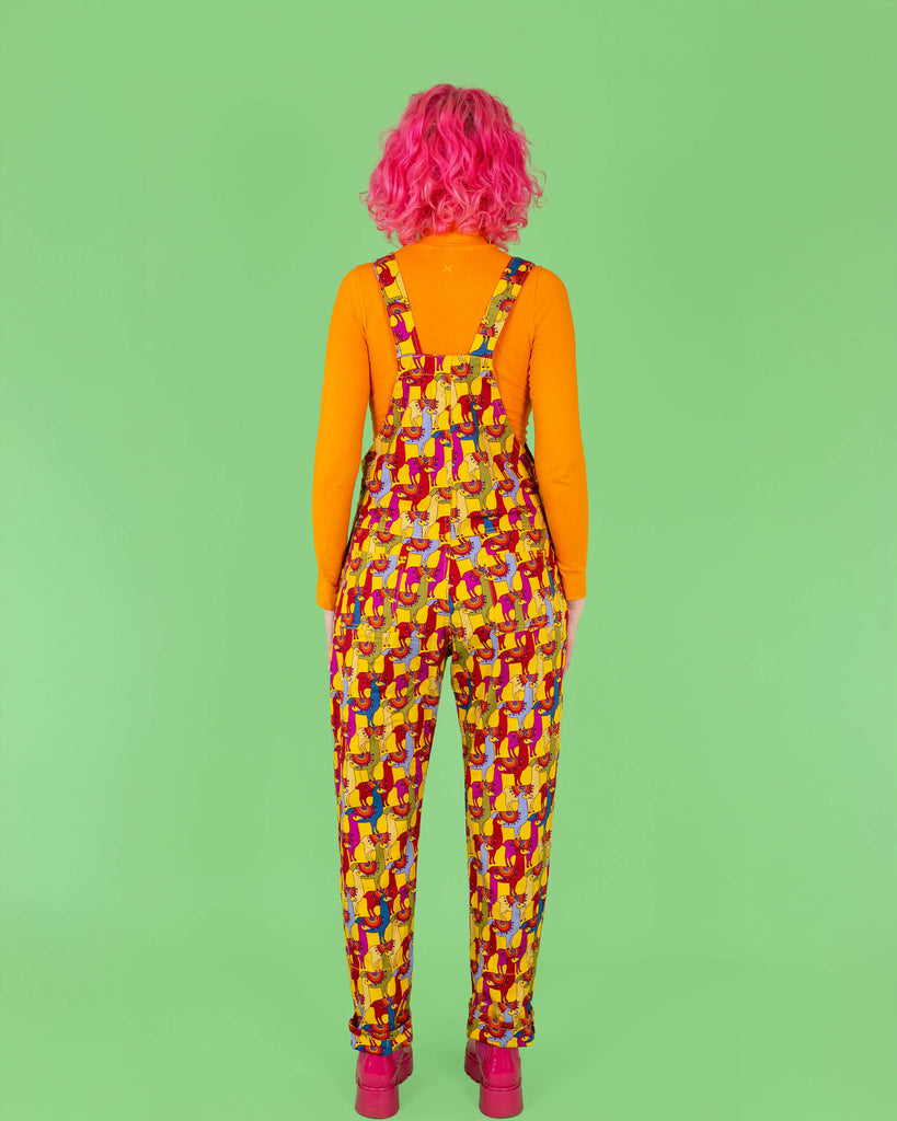 Back view of the model with pink hair in yellow dungarees covered in a vibrant llama print from Run & Fly. The dungarees feature bold, playful llamas in multiple colours, paired with an orange turtleneck and pink platform boots, creating a striking, fun look. Run & Fly is a small, ethical, slow fashion company.