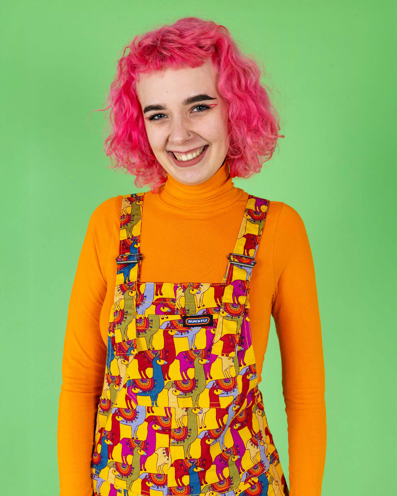 Close-up of a model with pink curly hair wearing yellow dungarees featuring a lively llama print in bold red, blue, and purple hues by Run & Fly. The bright orange turtleneck complements the fun design. The model's cheerful expression matches the playful and colourful vibe. Run & Fly is a small, ethical, slow fashion company.