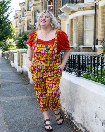 A model with curly blonde hair walks along a residential street, laughing, while wearing yellow dungarees with a colourful llama print from Run & Fly. The vibrant dungarees feature llamas in red, purple, and blue, and are paired with a red puff-sleeve top and sandals. The cheerful and casual outdoor setting complements the playful, fun vibe of the outfit. Run & Fly is a small, ethical, slow fashion company.