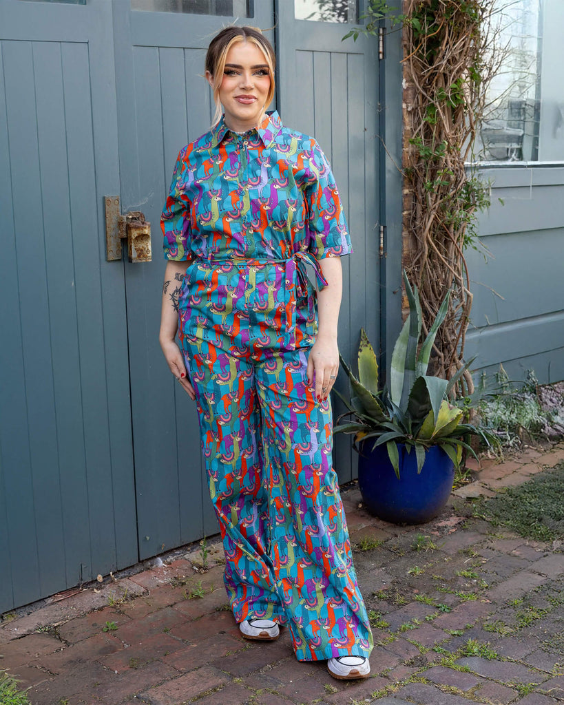 Model is wearing Party Llama Stretch Jumpsuit paired with trainers. The jumpsuit is a bright blue with llamas printed on in red, green, blue and purple. The jumpsuit is buttoned up to the collar and has a matching fabric belt tied round the front of the waist. Model is smiling at the camera.