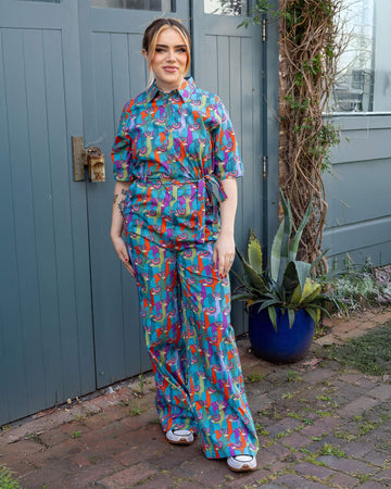 Model is wearing Party Llama Stretch Jumpsuit paired with trainers. The jumpsuit is a bright blue with llamas printed on in red, green, blue and purple. The jumpsuit is buttoned up to the collar and has a matching fabric belt tied round the front of the waist. Model is smiling at the camera.