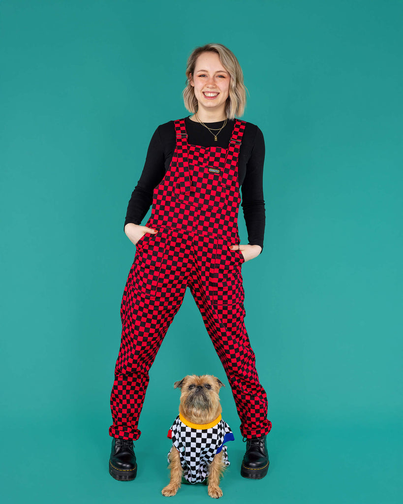Amy a blonde model with should length bobbed hair smiling at camera wearing black and magenta pink dungarees with Brian the Brussels Griffon dog in a black and white checkboard jumper sat between her legs. They are on a green background
