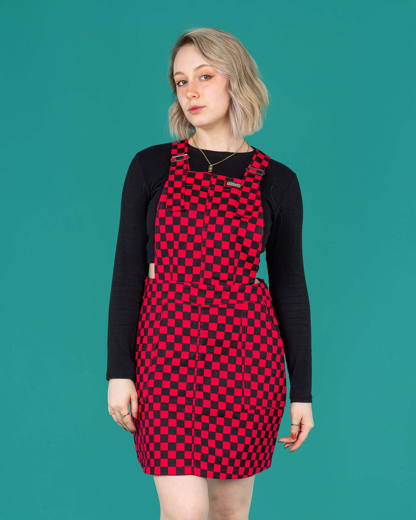 Amy a blonde white femme model with shoulder length hair is wearing a black long sleeved top and black boots with a pink and black pinafore dress stood against a green background