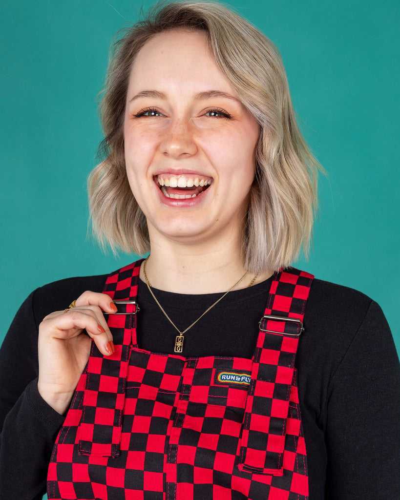Amy a blonde white femme model with shoulder length hair is wearing a black long sleeved top and black boots with a pink and black pinafore dress stood against a green background close up of Amy laughing