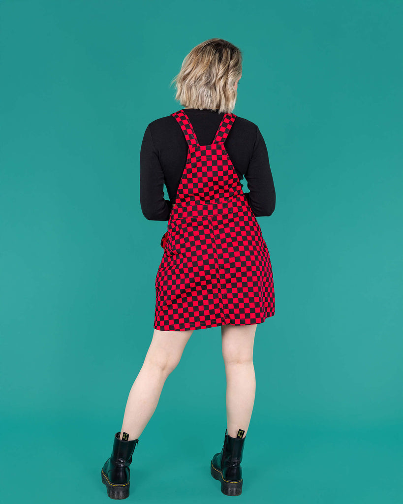 Amy a blonde white femme model with shoulder length hair is wearing a black long sleeved top and black boots with a pink and black pinafore dress stood against a green background she is facing the back