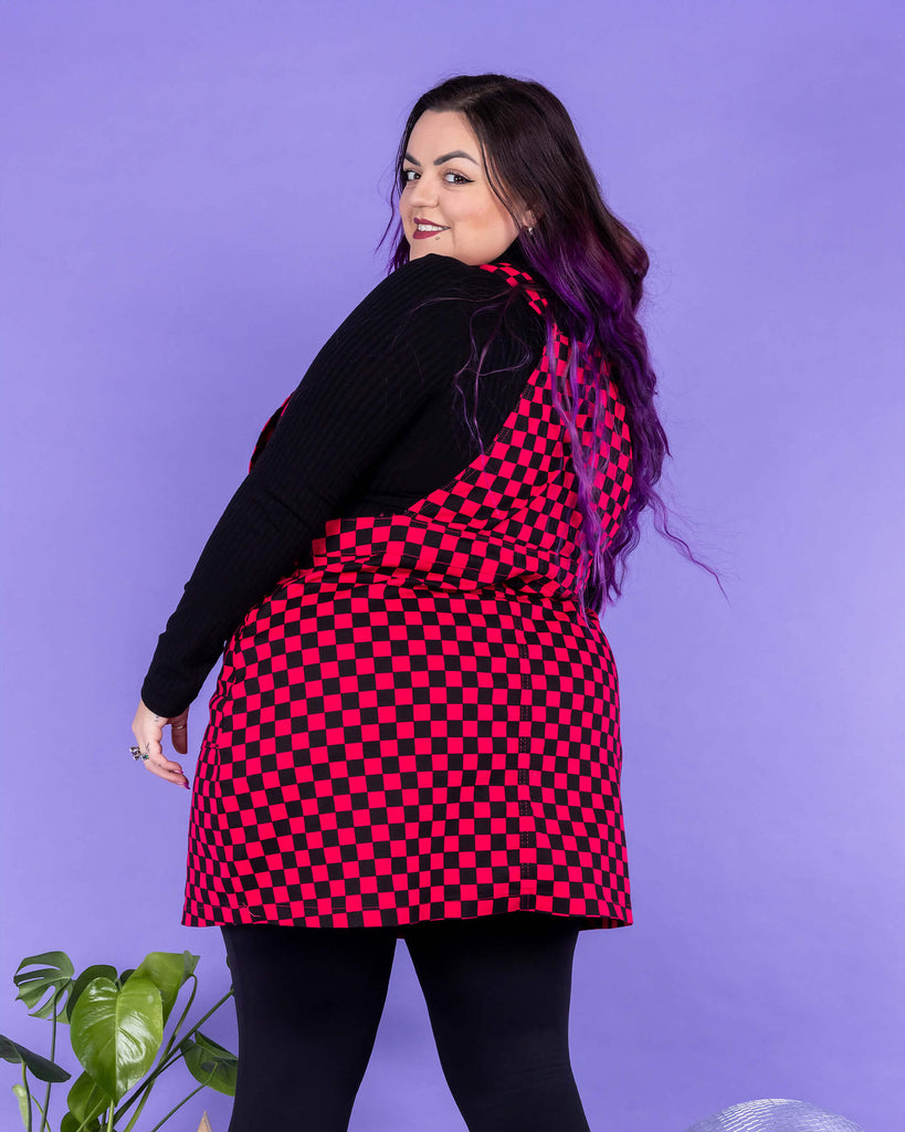 Luisa, a white femme model with purple hair, is wearing Black & Viva Magenta Checkerboard Stretch Twill Pinafore Dress with a long sleeve black turtle neck underneath and black tights and boots. Luisa is stood in front of a lilac background on a black fluffy rug amongst disco balls, a star shaped light and a plant with her back to the camera and is looking back over her shoulder smiling at the camera.