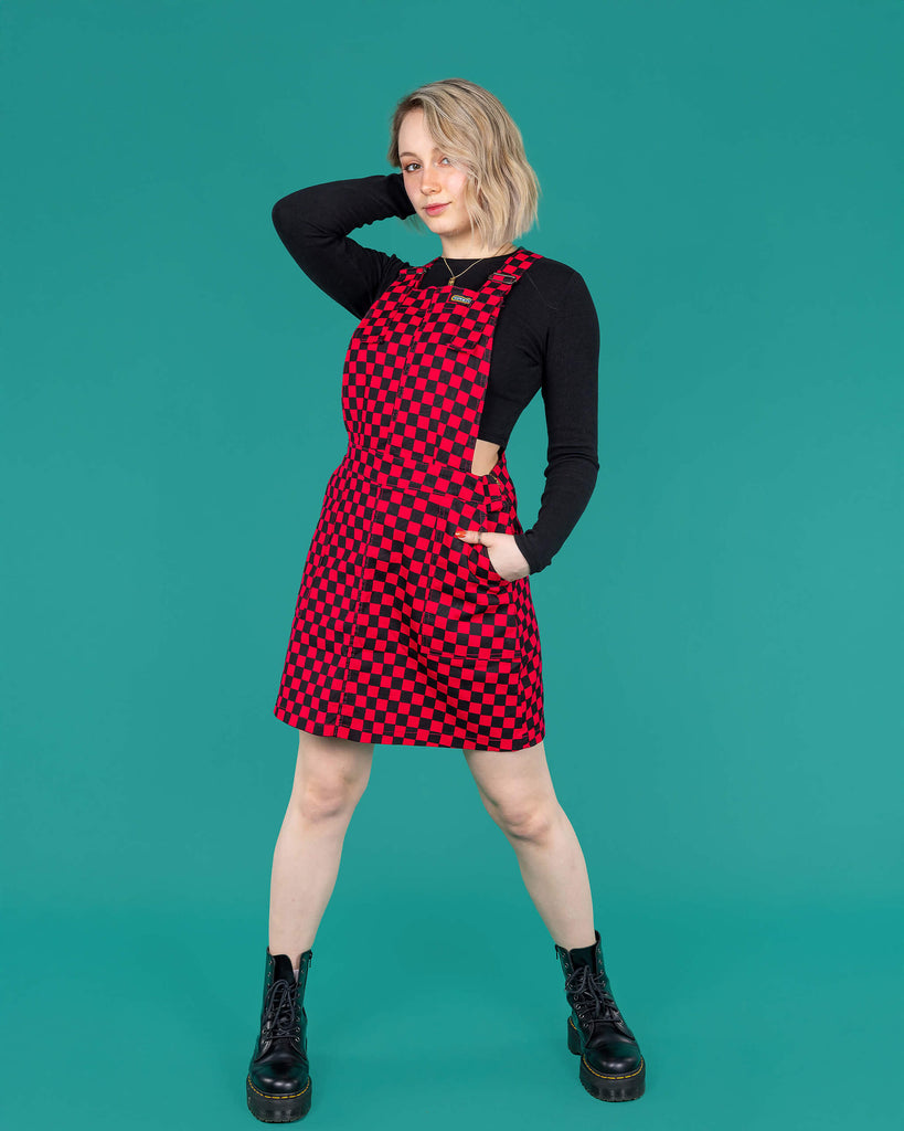 Amy a blonde white femme model with shoulder length hair is wearing a black long sleeved top and black boots with a pink and black pinafore dress stood against a green background.