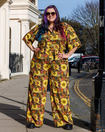 Model Luisa poses confidently in a Run & Fly jumpsuit designed by Jen James Design, adorned with sunflowers and fall-inspired flowers. She has colourful, wavy hair and sunglasses, standing in a sunny outdoor setting with her hands on her hips.