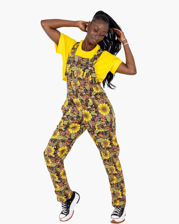 A model wearing Run & Fly’s Midnight Blues Stretch Twill Dungarees with a bold sunflower, mushroom, and foliage print. The dungarees are paired with a bright yellow T-shirt, highlighting the detailed floral design. This ethical slow fashion piece features adjustable straps and a front bib pocket, perfect for vibrant autumnal style. The model poses confidently, showcasing the relaxed fit and vibrant colours.