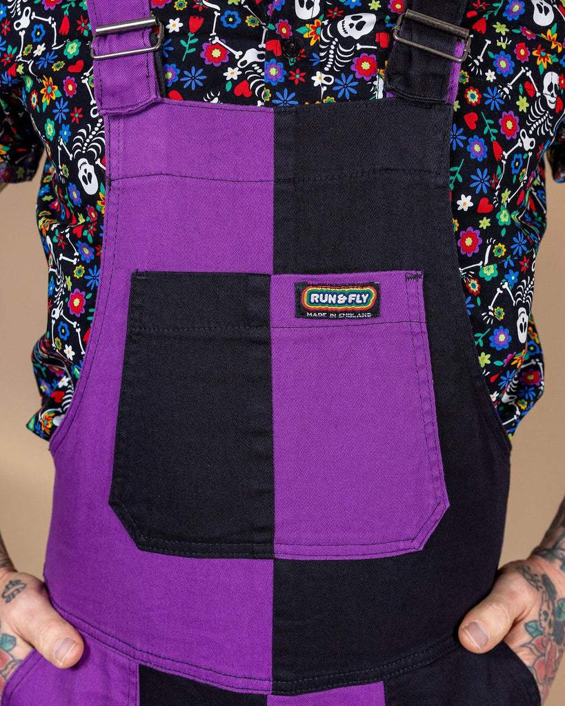 Close up of James wearing the boogie bones shirt and the purple and black mix n match two tone stretch twill dungarees in front of a beige background. The photo is cropped in on the chest to highlight the purple and black colourblock front pocket with the run and fly label at the top. The dungarees are made slowly and ethically in england.