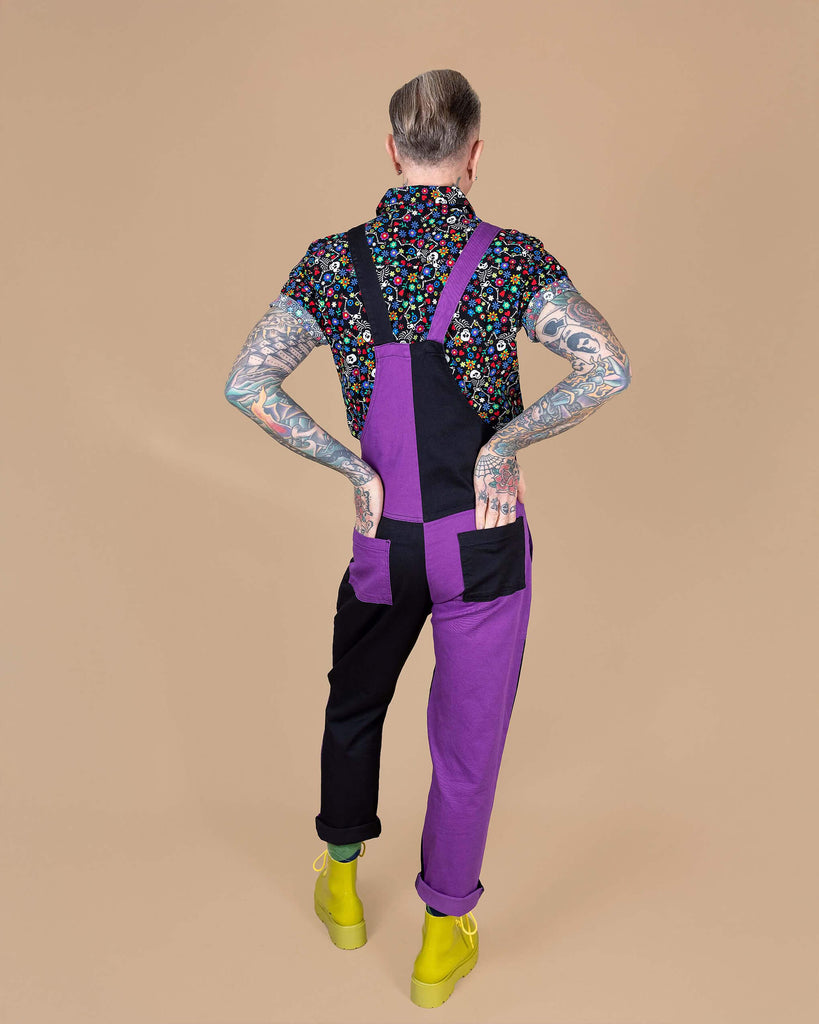 James is stood in front of a beige background wearing the boogie bones shirt and the purple and black mix n match two tone stretch twill dungarees and yellow boots. He is facing away from the camera leaning more on one leg with both hands resting in the back dungaree pockets. The dungarees are made slowly and ethically in england.