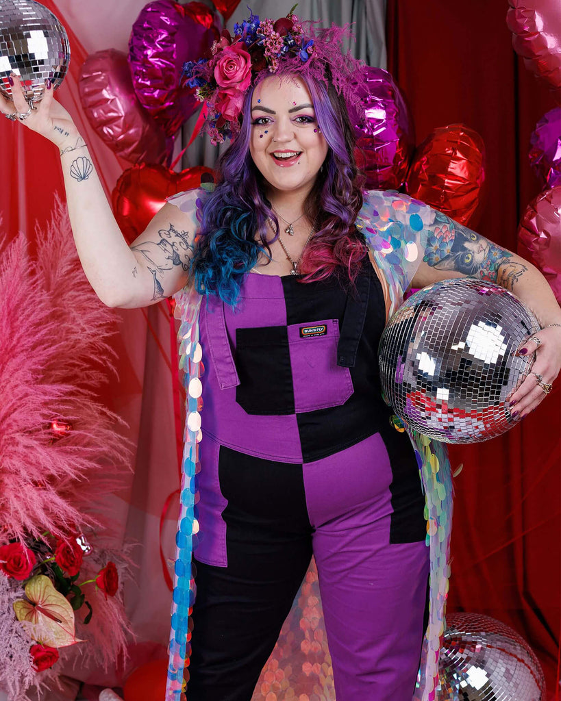 Luisa, a white femme model with purple, blue and pink in their hair is stood in a studio in Hove with a red backdrop with heart shaped balloons and pink feathers and rose petals on the floor. They are wearing Purple and Black Mix n Match Two Tone Stretch Twill Dungarees with a sequin shawl and floral headdress. Luisa is posing holding disco balls and facing the camera. 