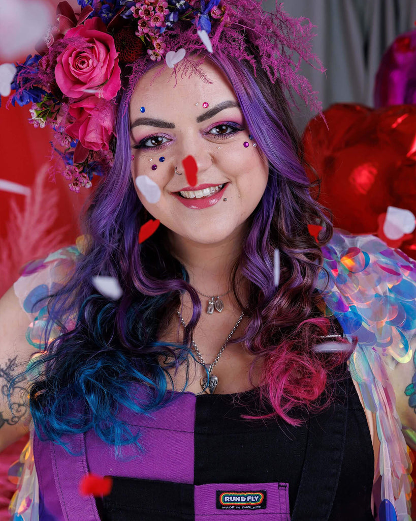 Luisa is posing in a photography studio in Hove with a floral headdress, and gems on their face with pink eye makeup. She is wearing Purple and Black Mix n Match Two Tone Stretch Twill Dungarees and is smiling with the photo cropped at the chest.