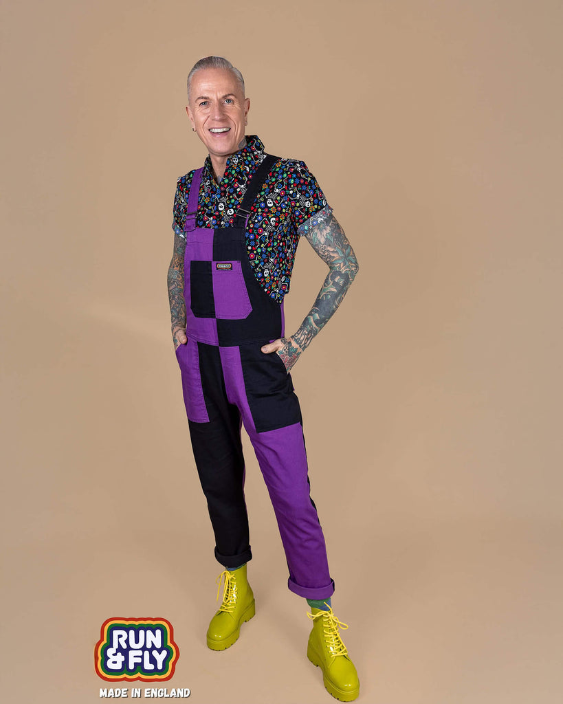 James is stood in front of a beige background wearing the boogie bones shirt and the purple and black mix n match two tone stretch twill dungarees and yellow boots. He is facing forward stood leaning back on one leg with both hands in the dungaree pockets whilst smiling. In the bottom left corner there's a made in England logo.