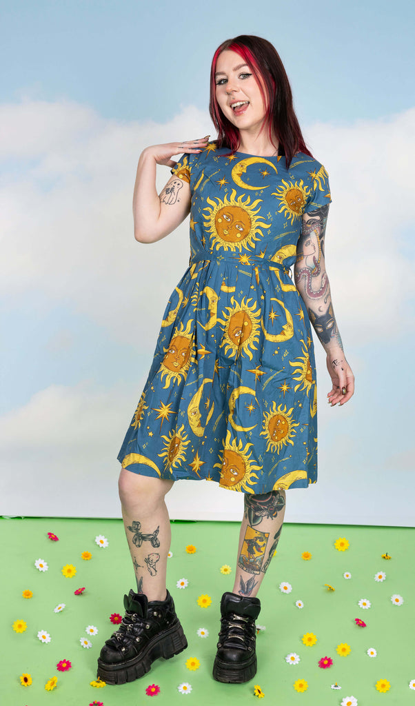Florence is stood in a photography studio in Hove in front of blue sky backdrop on a green floor with flowers scattered wearing Celestial Sun and Moon Stretch Belted Tea Dress with Pockets with chunky black boots. The dress print is a denim blue colour with gold and yellow retro style suns and moons with faces, shooting stars, stars and sparkles. Florence is smiling at the camera and posing with one hand resting on her shoulder and one leg slightly bent outwards.