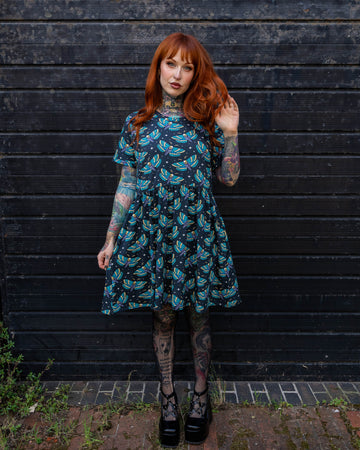 A model, Erin Porter, with long red hair is standing against a black wooden wall, wearing a Run & Fly dress with a colourful death's-head hawkmoth print. The dress features bright blue and yellow moths on a dark background. The model's tattooed arms and legs give the look an edgy, alternative style. Run & Fly is a small, ethical, slow fashion company.