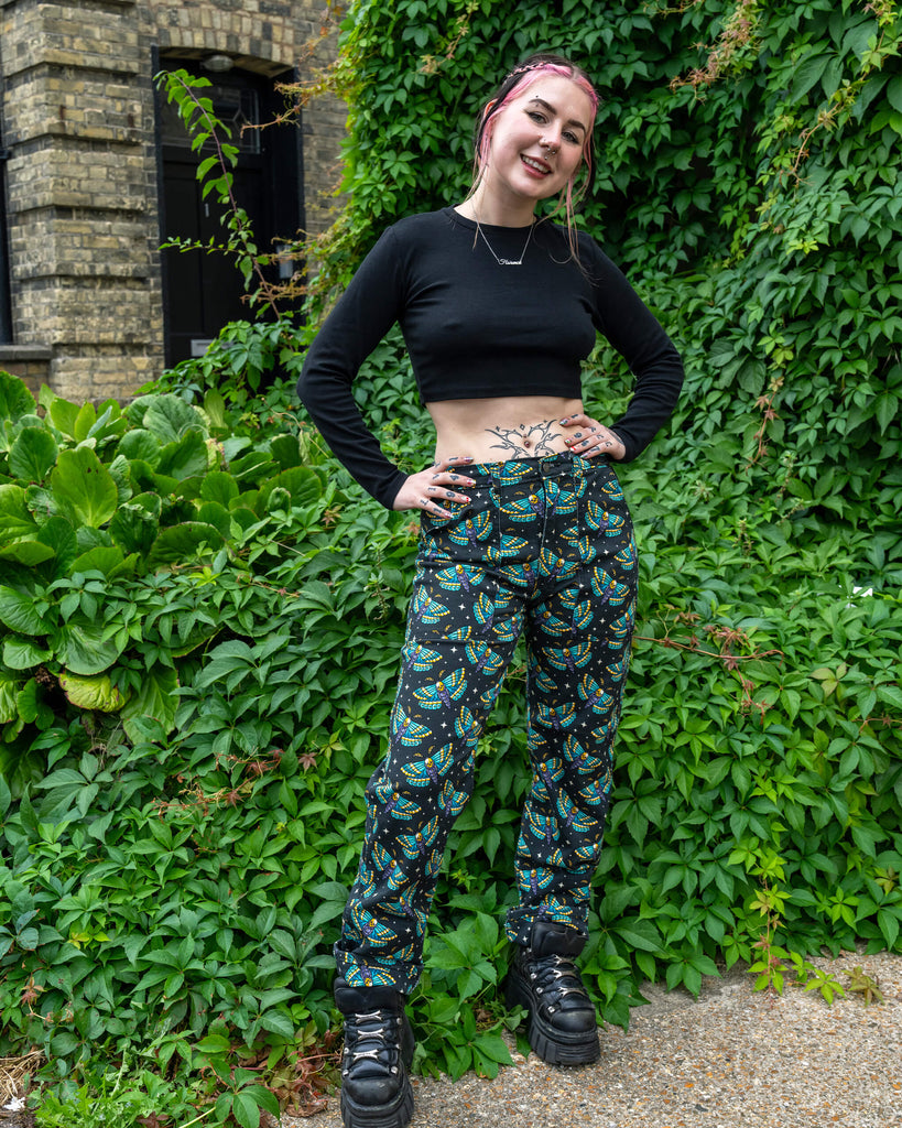A person models a pair of moth-print trousers, featuring a detailed blue, yellow, and purple design against a black background. They are paired with a black crop top and platform shoes, highlighting Run & Fly’s ethical and alternative fashion.