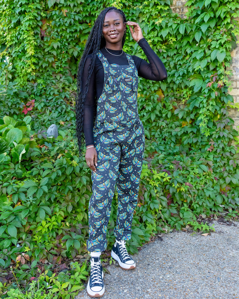 A model is stood outside in hove by greenery wearing Halloween: Death Head Moth Stretch Twill Dungarees with a long sleeved mesh top underneath and black trainers. The dungarees have a dark base with an all over print of blue and yellow death head moths amongst star bursts. She is facing the camera leaning slightly to one side with her hands resting in the pockets of the dungarees. 