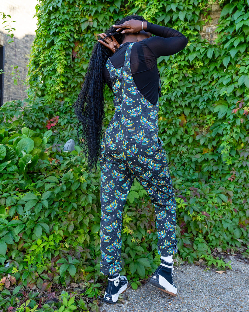 A model is stood outside in hove by greenery wearing Halloween: Death Head Moth Stretch Twill Dungarees with a long sleeved mesh top underneath and black trainers. The dungarees have a dark base with an all over print of blue and yellow death head moths amongst star bursts. She is stood with her back to the camera with her long hair swishing behind her. 