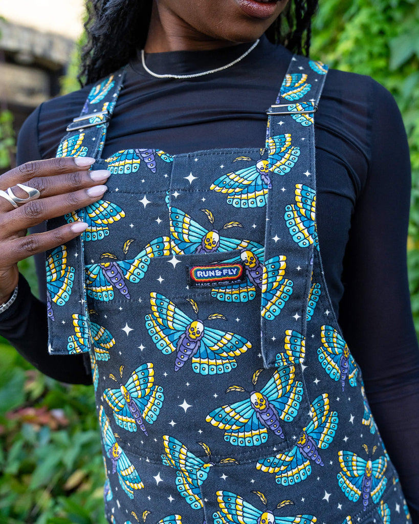 close up of model wearing wearing Halloween: Death Head Moth Stretch Twill Dungarees. The dungarees have a dark base with an all over print of blue and yellow death head moths amongst star bursts. The close up shows the Run & Fly logo on the bib of the dungarees