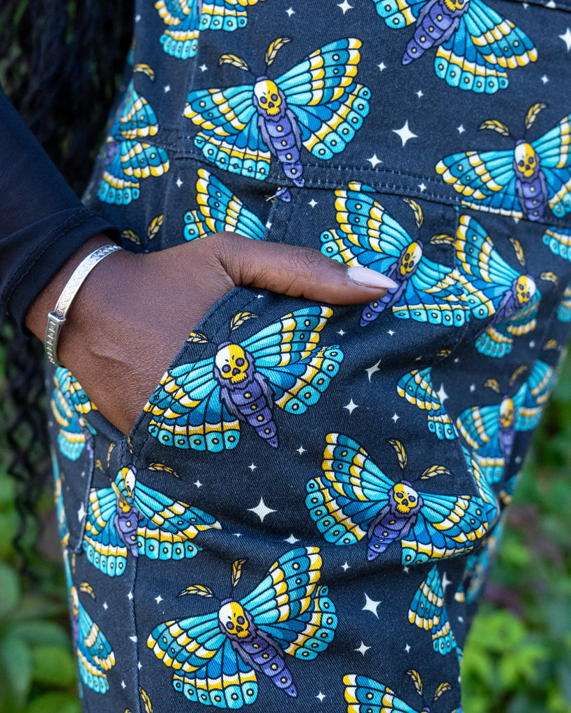 close up of model's hand in the pocket of the Halloween: Death Head Moth Stretch Twill Dungarees. The dungarees have a dark base with an all over print of blue and yellow death head moths amongst star bursts. 