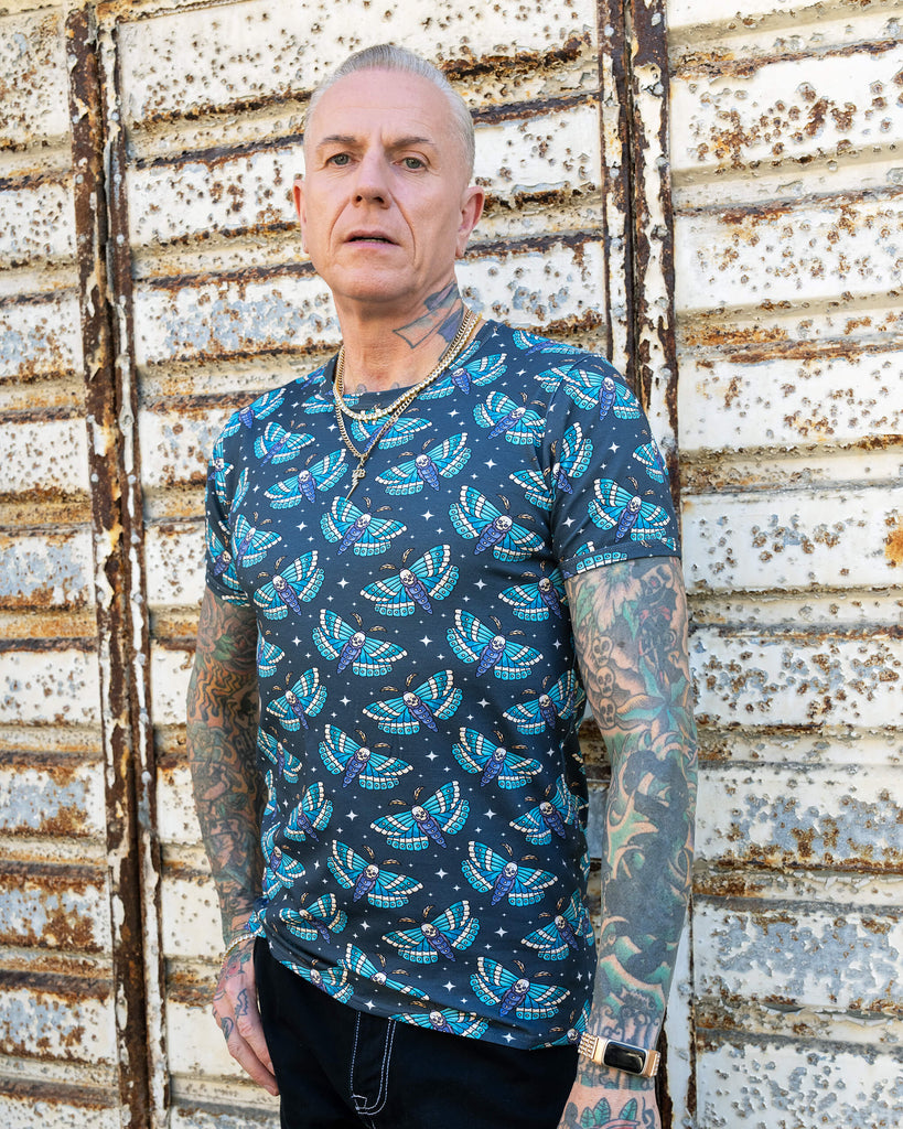 James stands confidently, facing the camera, showcasing the front of the “Death Head Moth” t-shirt. The intricate blue moth pattern with skulls pops against the dark background. His tattoos and jewellery add to the tough, stylish aesthetic.