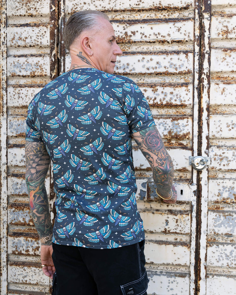 James, with tattooed arms, models a t-shirt featuring Run & Fly’s “Death Head Moth” print. The back of the shirt displays a repeating pattern of blue moths with skull details against a dark background. The distressed metal door adds an edgy contrast to the design.