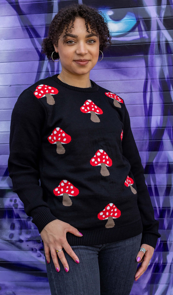 Georgia is stood in front of a purple graffiti wall wearing Mushroom Print Black Jumper with jeans. The black long sleeved jumper has an all over red and white mushroom print on the torso. The model is posing facing the camera. 