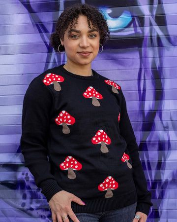 Georgia is stood in front of a purple graffiti wall wearing Mushroom Print Black Jumper with jeans. The black long sleeved jumper has an all over red and white mushroom print on the torso. The model is posing facing the camera. 