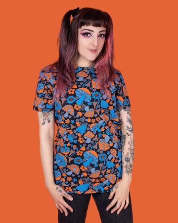 Sarah is stood in front of an orange background wearing the Magic Mushroom Short Sleeve Tee with black stonewash denim flares. She is facing forward with both hands resting on her hips whilst smiling to camera. The magic mushroom print is a black base tshirt with blue and neon orange toadstool mushrooms and daisy shape flowers all over.