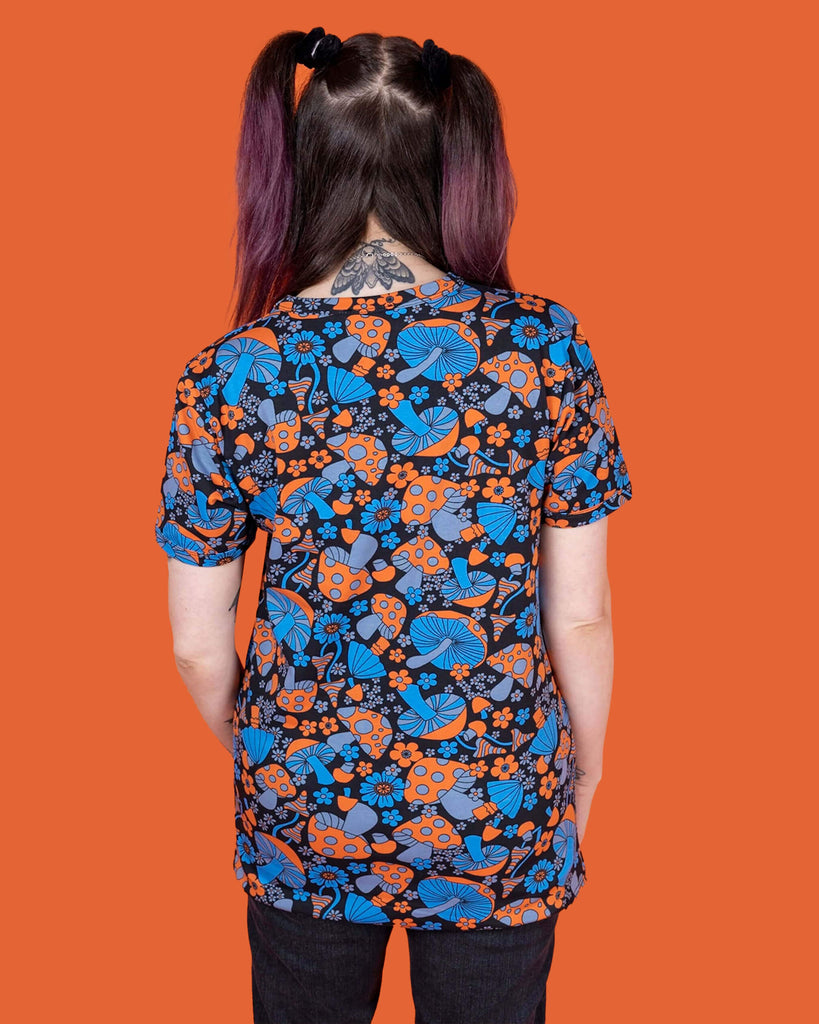 Sarah is stood in front of an orange background wearing the Magic Mushroom Short Sleeve Tee with black stonewash denim flares. She is facing away from the camera with both hands resting by her sides. The magic mushroom print is a black base tshirt with blue and neon orange toadstool mushrooms and daisy shape flowers all over.