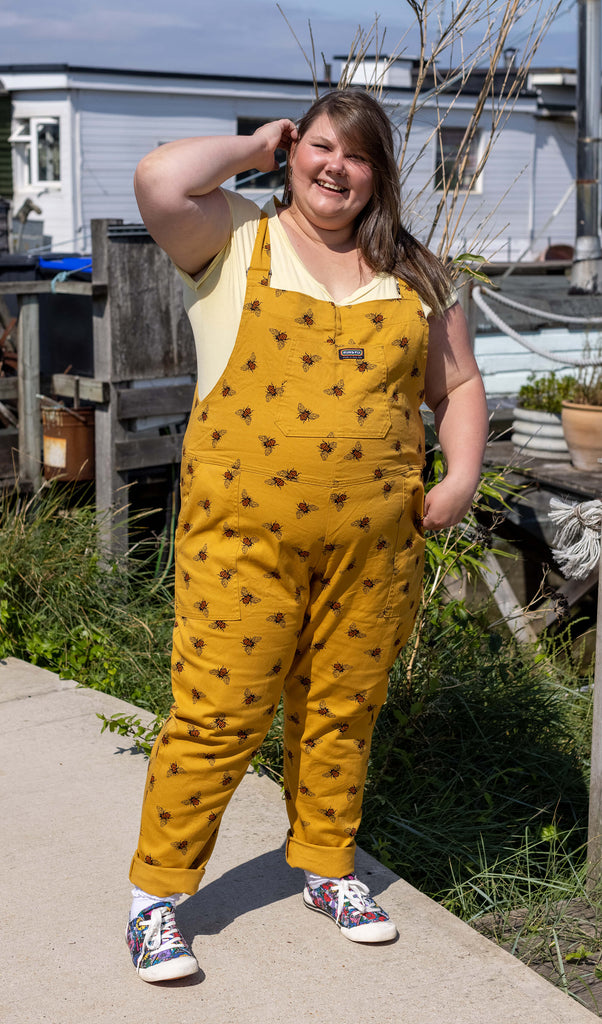 Full length photo of Natalie wearing the Bees Knees Gold Stretch Twill Dungarees with a yellow tee and canvas trainers. Her hand is pushing her hair out of her face and she's smiling at the camera.