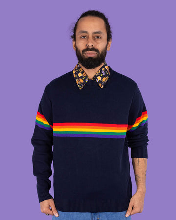 Richard, a hispanic male model with dark hair in a bun and a beard, is stood in a photography studio in Hove in front of a lilac backdrop wearing Rainbow Navy Jumper with Highland Cows Short Sleeve Shirt and blue jeans. Richard is facing the camera and is pulling the jumper down slightly with his hands. The photo is cropped at the thighs. 