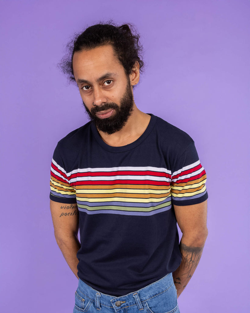 Richard, a hispanic male model with dark hair in a bun and a beard, is stood in a photography studio in Hove in front of a lilac backdrop wearing Navy Stripe Tee with blue jeans. Richard is posing toward the camera leaning forward slightly with his arms behind his back. The photo is cropped at the waist.