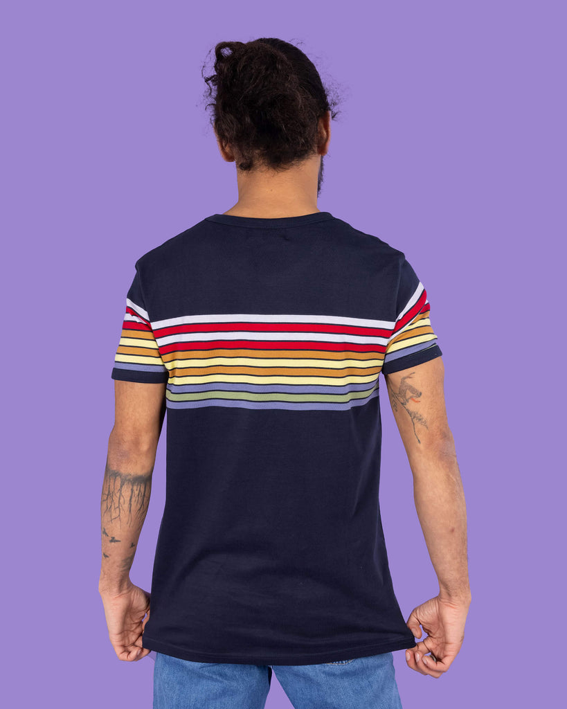 Richard, a hispanic male model with dark hair in a bun and a beard, is stood in a photography studio in Hove in front of a lilac backdrop wearing Navy Stripe Tee with blue jeans. Richard is posing with his back to the camera and is pulling down the t shirt slightly. The photo is cropped at the thighs.
