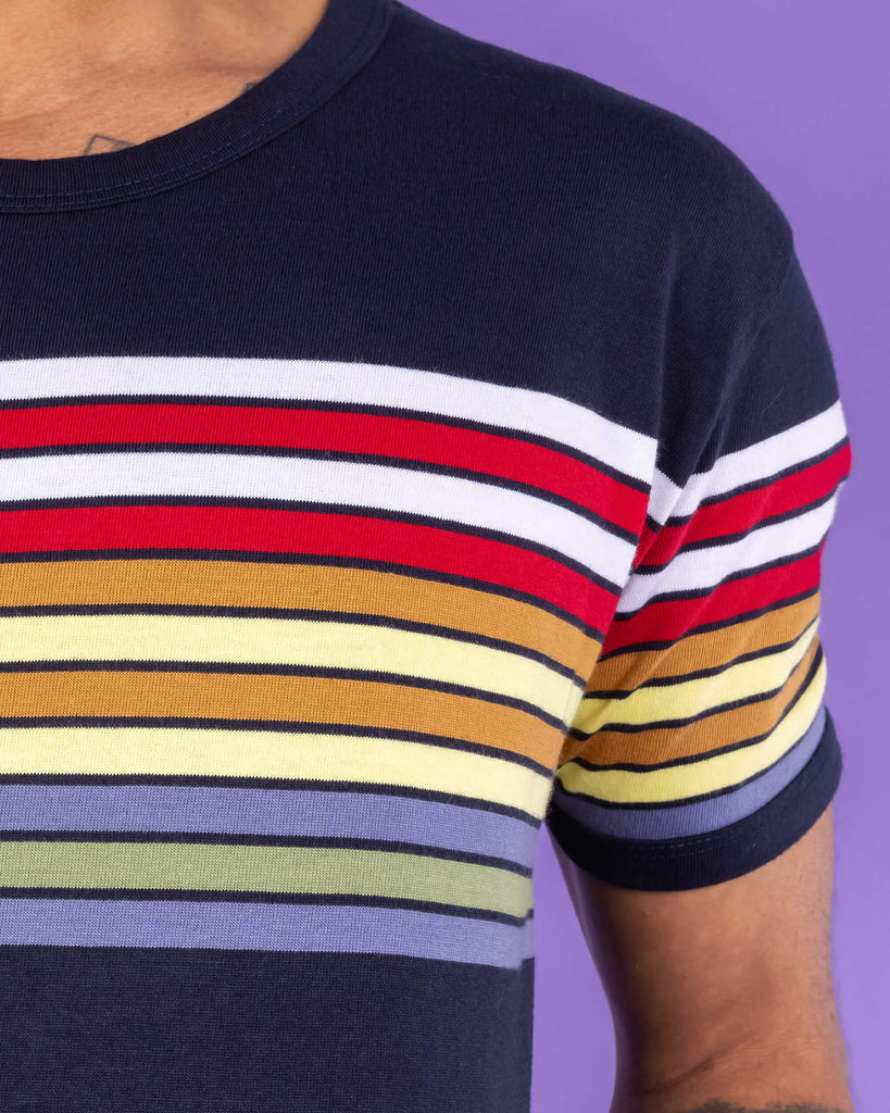 Close up of the Navy Stripe Tee. The navy tee has horizontal stripes across the chest in white, red, orange, yellow, green and blue. 