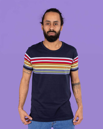 Richard, a hispanic male model with dark hair in a bun and a beard, is stood in a photography studio in Hove in front of a lilac backdrop wearing Navy Stripe Tee with blue jeans. Richard is posing toward the camera, slightly pulling down the t shirt with his hands. The photo is cropped at the thighs.