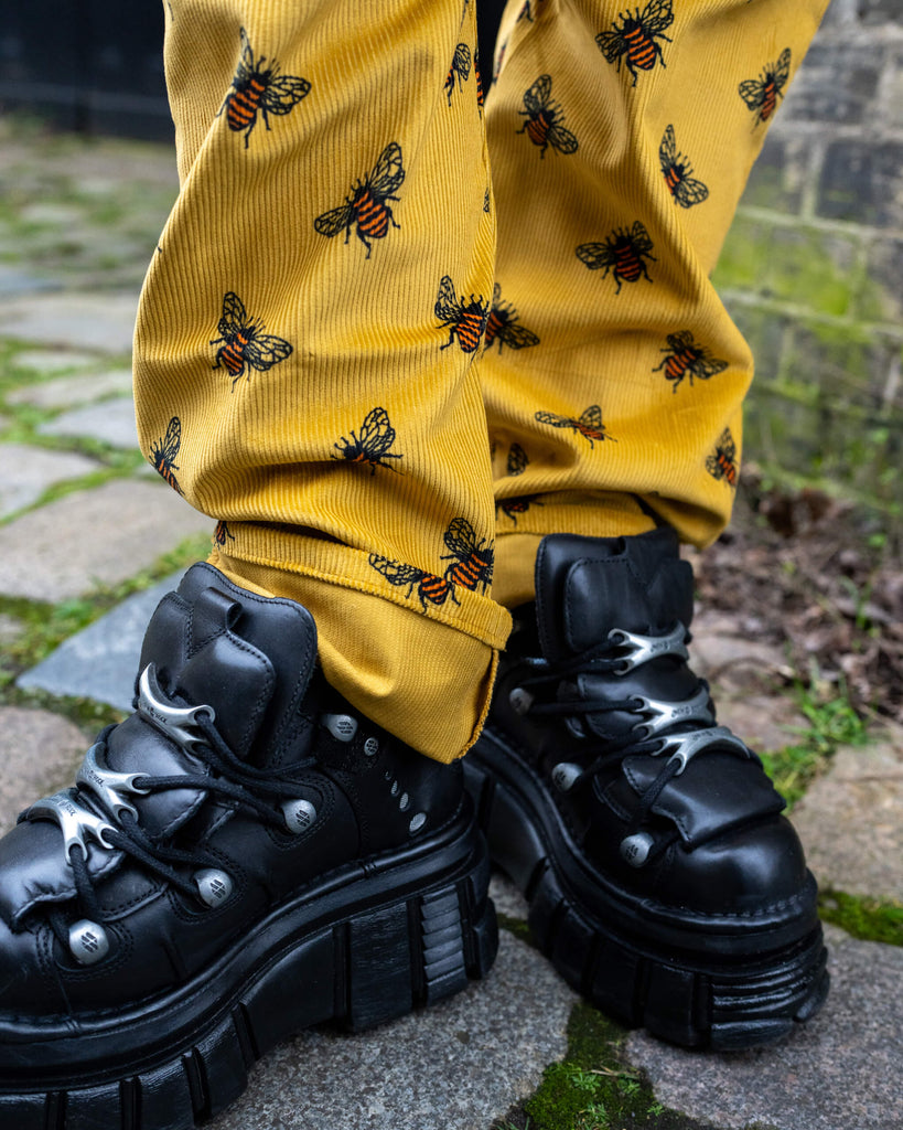 A detailed shot of the hem of Run & Fly’s Gold Bee Corduroy DungaBee Dungarees, showcasing the mustard yellow corduroy fabric and embroidered bee print. The model wears chunky black platform boots with silver hardware, adding an alternative twist to the vintage-inspired style.